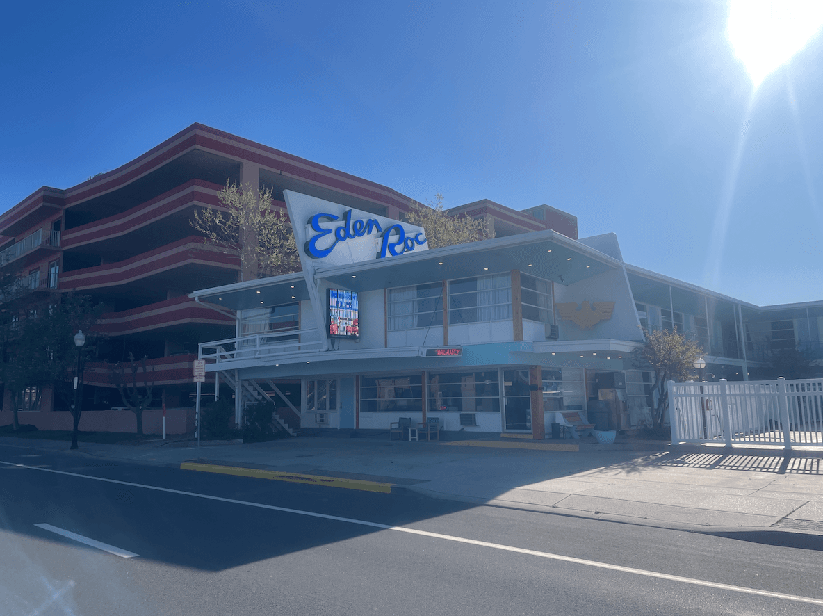 Front view of Eden Roc, a retro-style hotel/restaurant with a blue sign, glass windows, and outdoor seating on a sunny day.