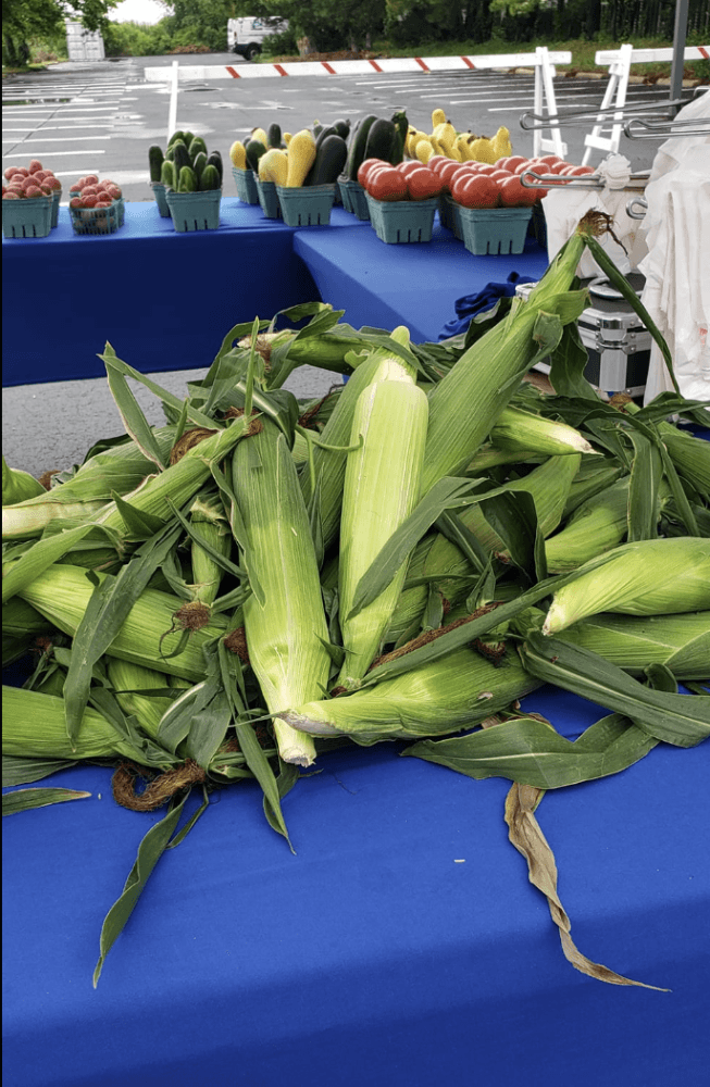 Fresh ears of corn with green husks piled on a blue market table at an outdoor market, with baskets of vegetables in the background.