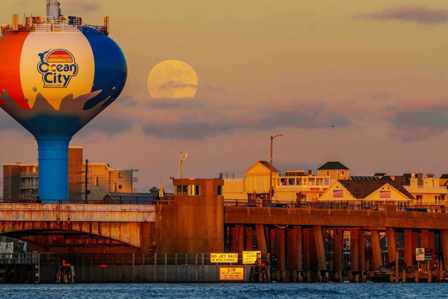 Large blue, red, and white water tower labeled Ocean City Maryland overlooking a pier at sunset with a rising moon in the sky.