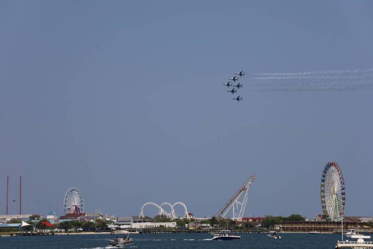 Five military jets fly in tight formation over a coastal amusement park with a ferris wheel and roller coasters, leaving white contrails across a clear blue sky.