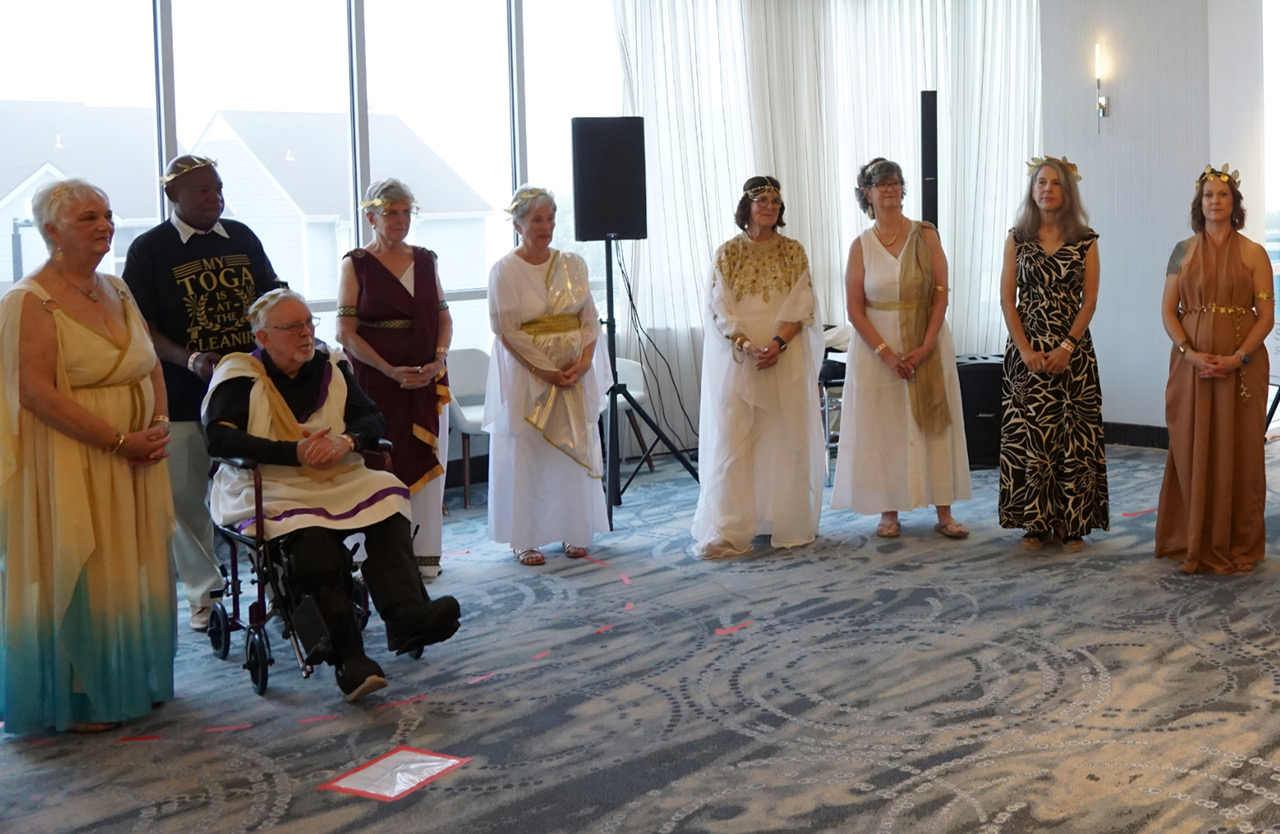 Group of nine people in Greek robes with laurel crowns standing in a bright event space; a man in a wheelchair is center-left.