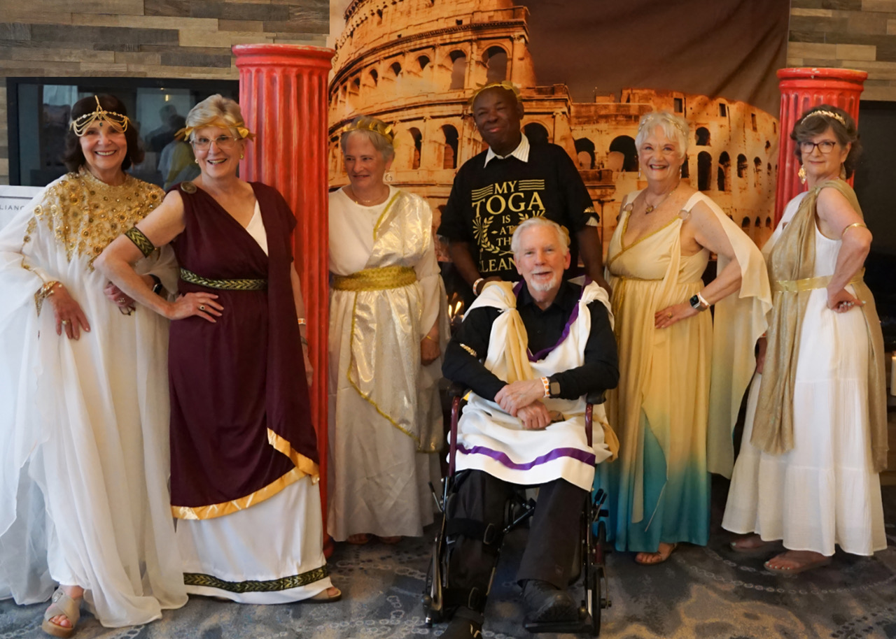 Group of adults in gold-trimmed ancient robes posing with a smiling man in a wheelchair in front of a Roman panorama backdrop.