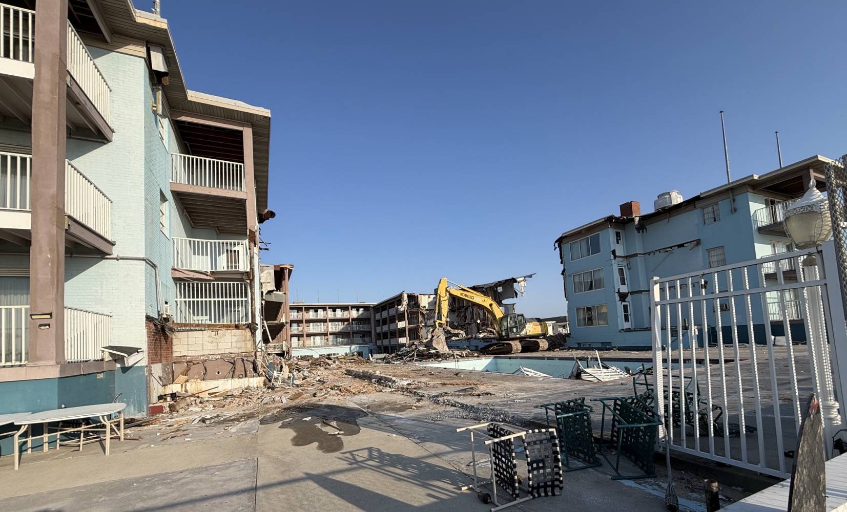 Demolition scene: a yellow excavator tearing down a partially collapsed building amid rubble and blue apartment blocks. ready to proceed with debris removal