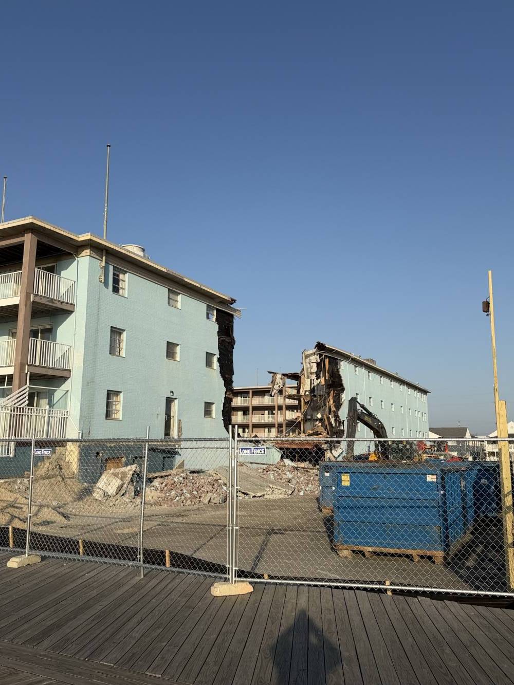 Construction site behind a chain-link fence with a blue dumpster and piles of rubble near a mint-blue building, on a wooden boardwalk