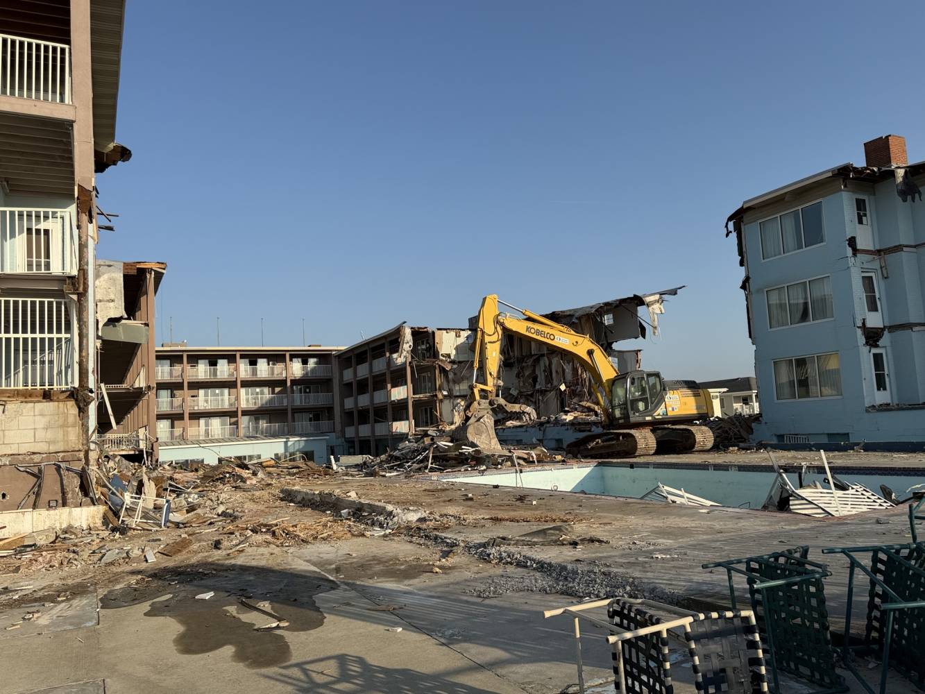 Yellow excavator demolishing a partially collapsed apartment building, rubble scattered across the site.