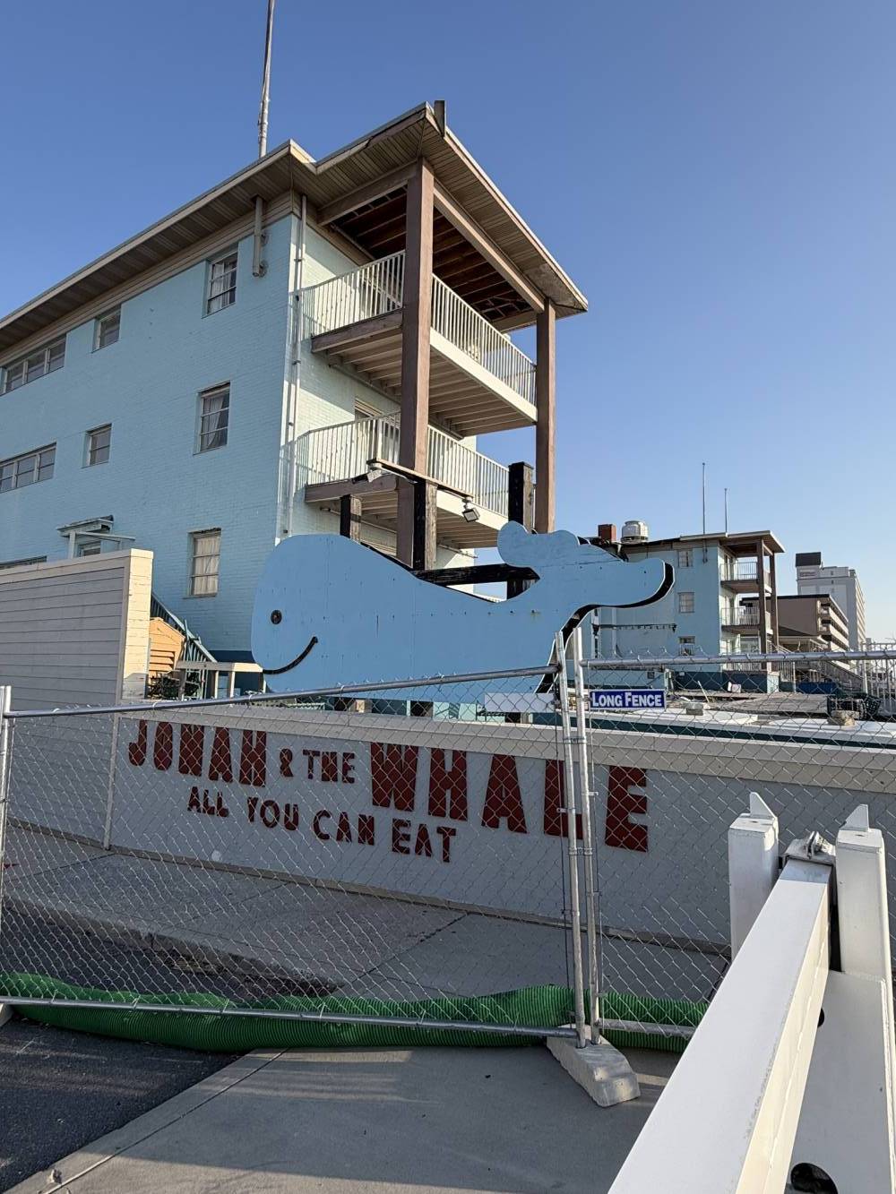 Blue whale sculpture attached to a chain-link fence beside a pale blue building, with a 'Long Fence' sign in view.