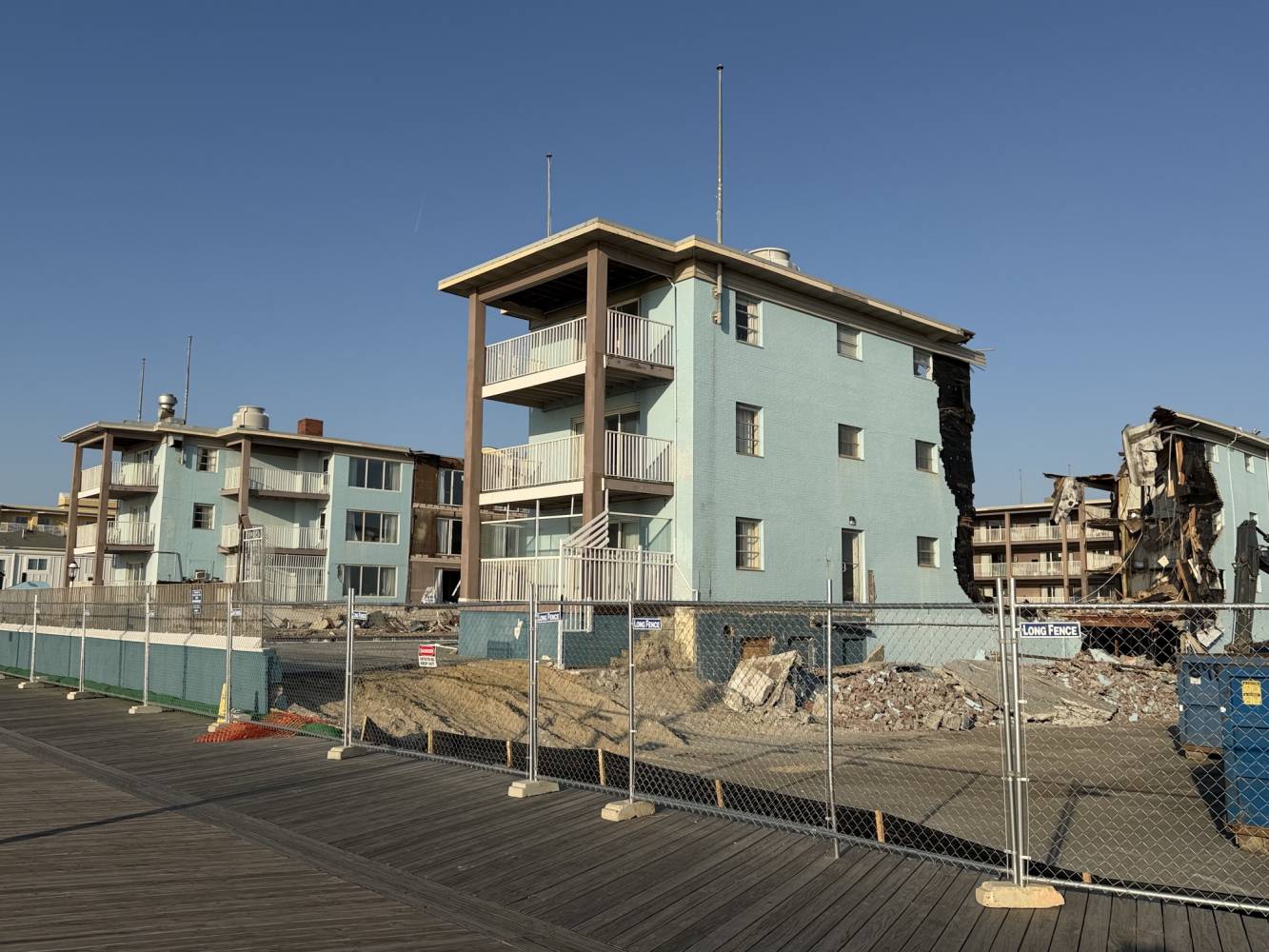 Blue apartment building with a damaged side and rubble behind a chain-link fence on a construction site, under a clear sky