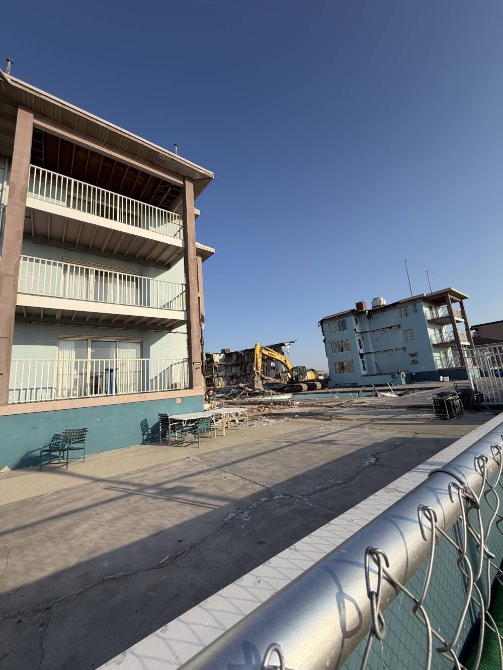 Construction site along a waterfront: a yellow excavator works beside a partially demolished building, with a chain-link fence in the foreground and a clear blue sky.