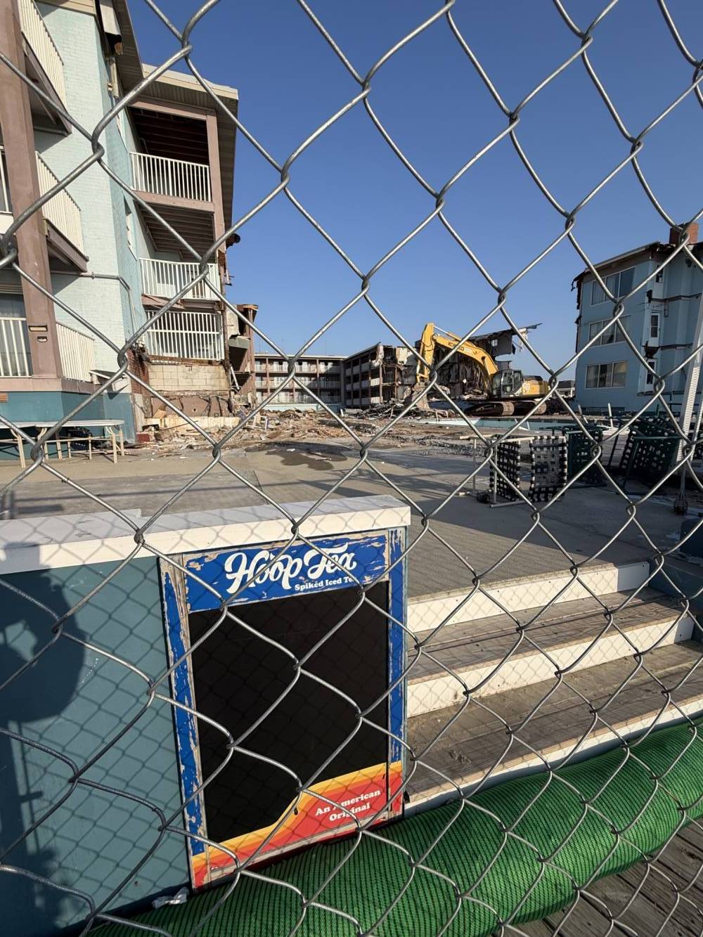 Construction site behind a chain-link fence with a yellow excavator, stacked pallets, and a building under construction against a blue sky.