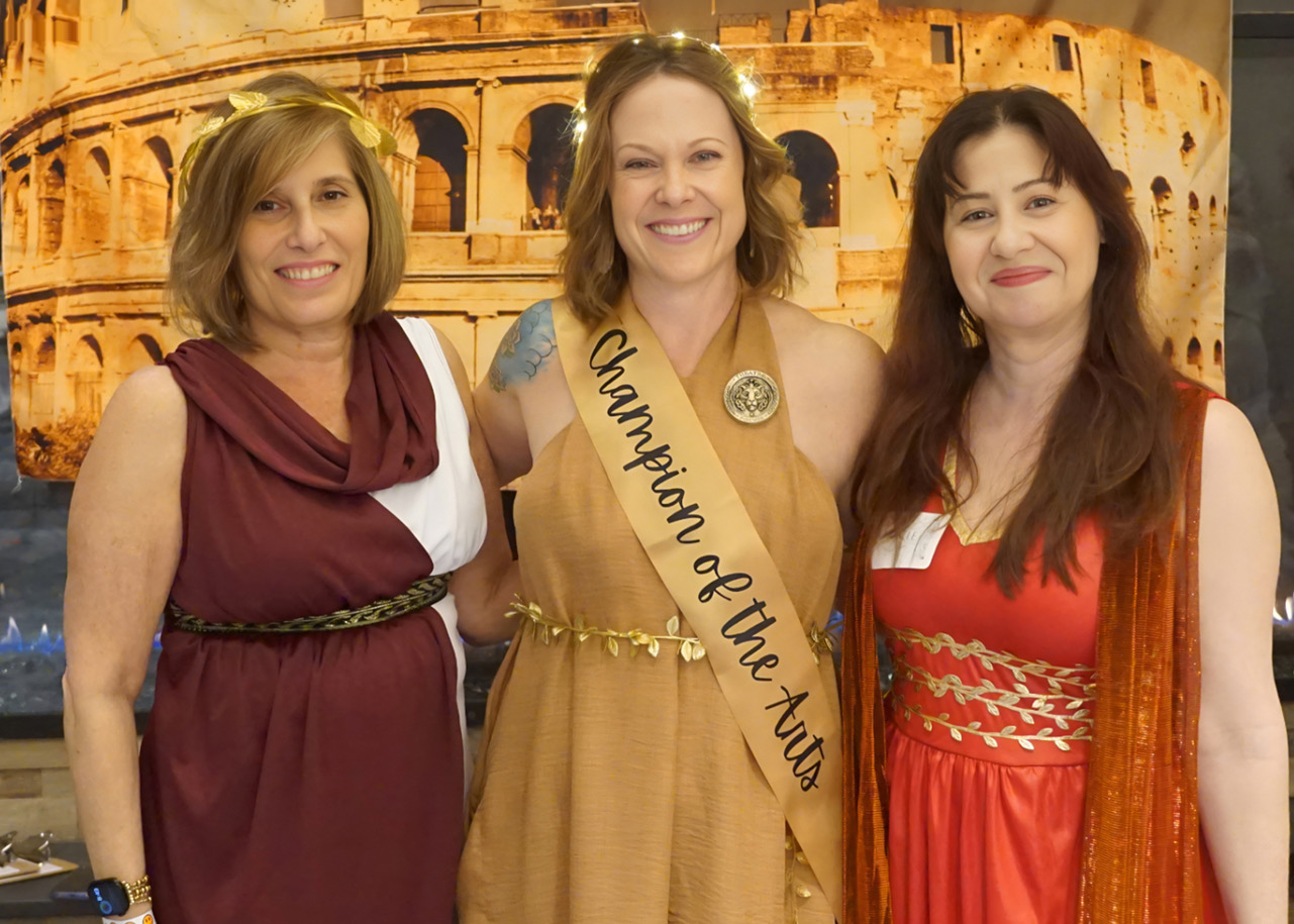Three women posing together at an awards event; center wears a tan dress with a sash reading 'Champion of the Arts' and a medal, with a Colosseum backdrop.