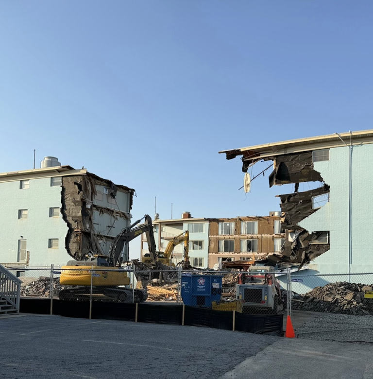 Construction site: a yellow excavator demolishes a pale-blue apartment building, with collapsed walls and rubble behind a chain-link fence under a clear blue sky.