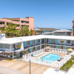 A retro seaside motel with a rectangular pool, white fencing, and pastel blue balconies, facing the ocean.