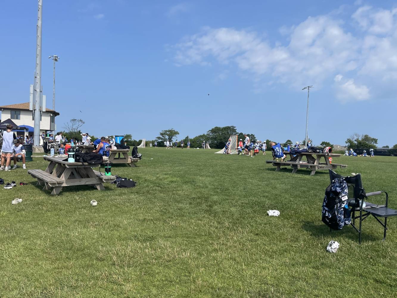 Youth lacrosse practice on a sunny day, players in white shirts near goal nets with picnic tables and gear on the grass.