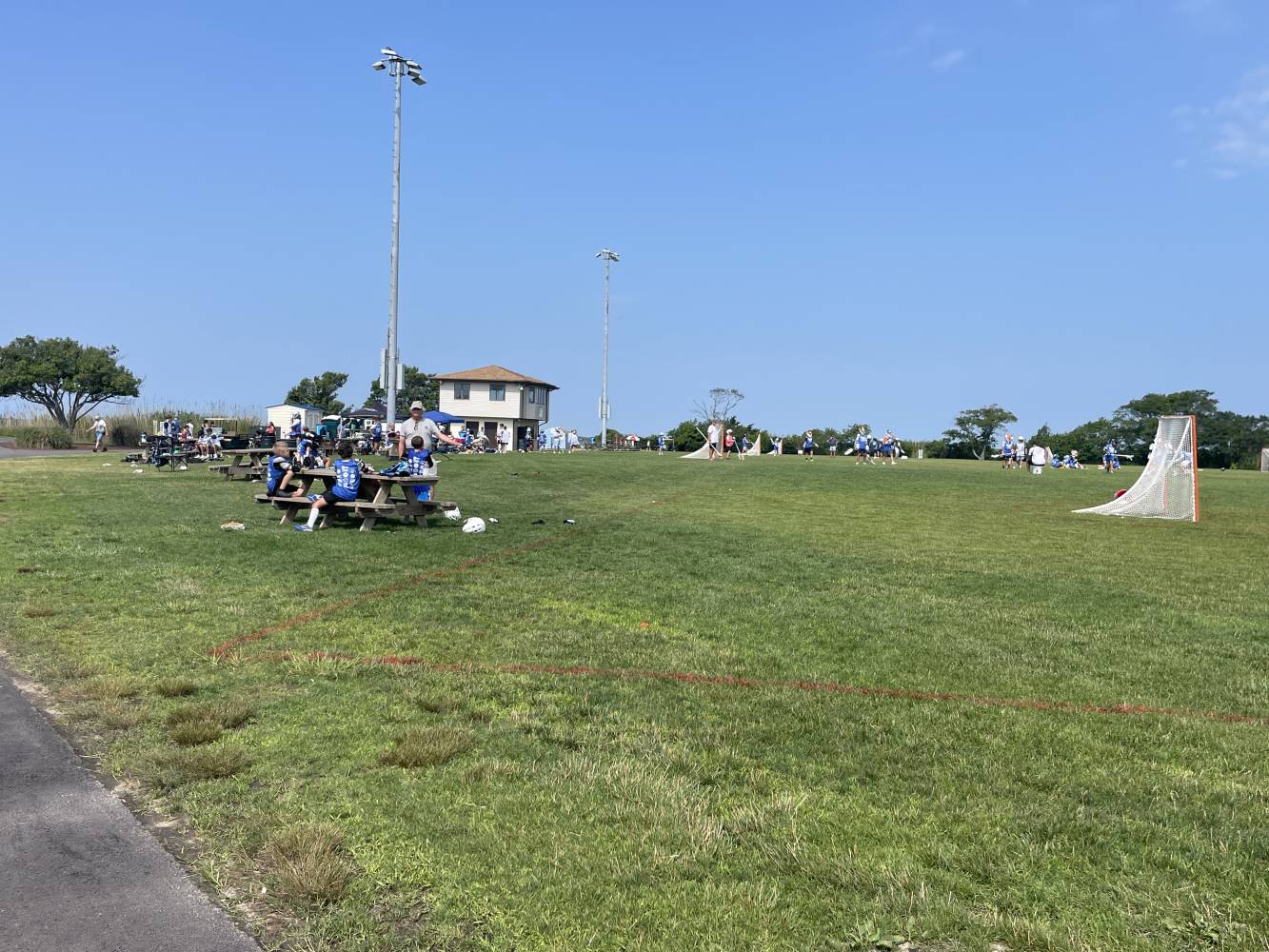 Sunlit park with groups of players in blue uniforms near picnic tables and a lacrosse goal on the grassy field.